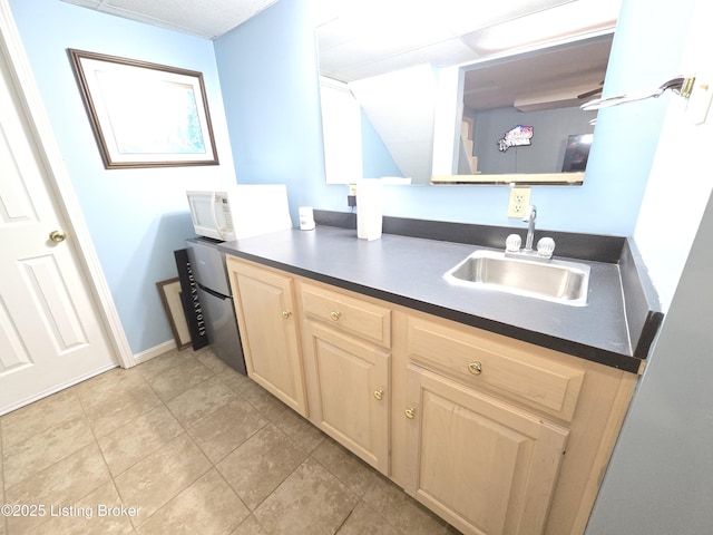 kitchen featuring dark countertops, light brown cabinetry, white microwave, light tile patterned flooring, and a sink