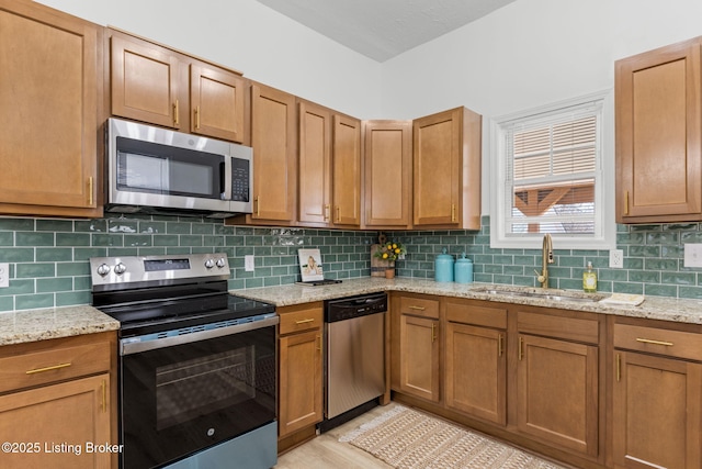 kitchen featuring a sink, tasteful backsplash, appliances with stainless steel finishes, brown cabinetry, and light stone countertops