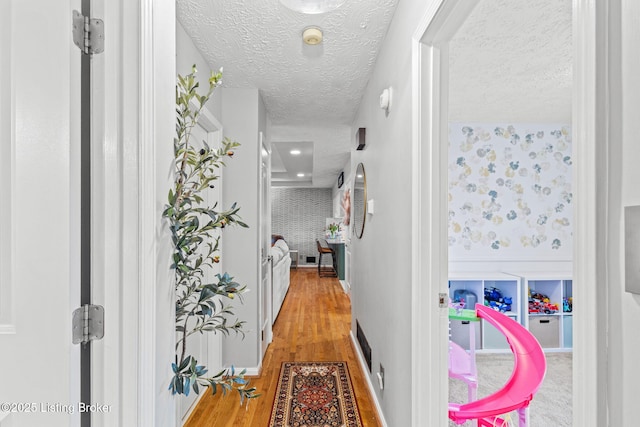 hall with wood finished floors, a textured ceiling, baseboards, and wallpapered walls