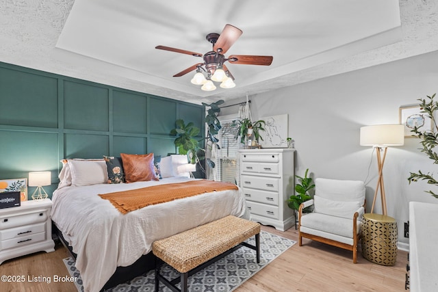 bedroom featuring ceiling fan, a tray ceiling, a decorative wall, and wood finished floors