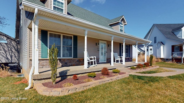 view of front of property with stone siding, covered porch, a shingled roof, and a front yard