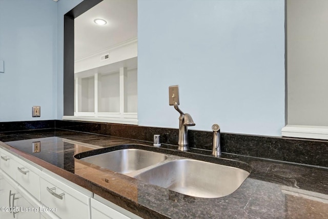 kitchen featuring a sink, visible vents, white cabinets, ornamental molding, and dark stone counters