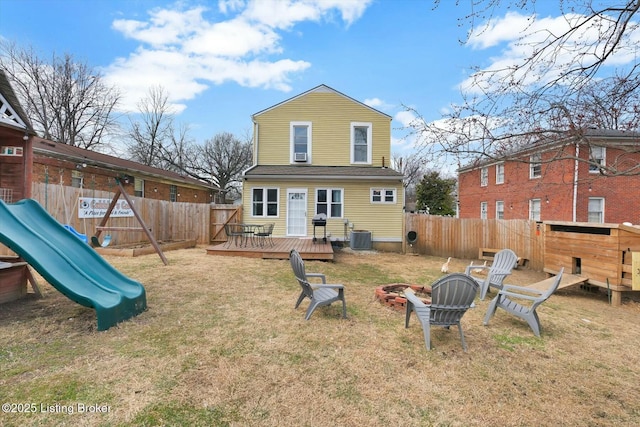 back of property with a wooden deck, a playground, a fenced backyard, and an outdoor fire pit