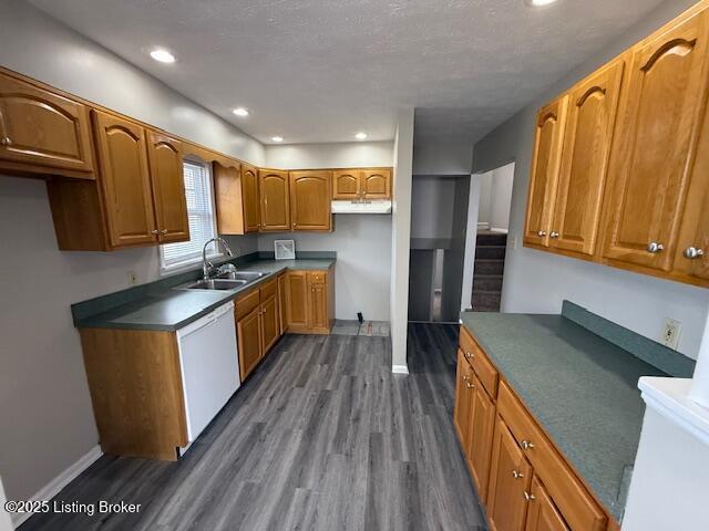 kitchen with brown cabinetry, dark wood finished floors, white dishwasher, under cabinet range hood, and a sink