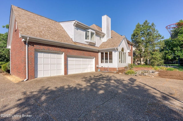 view of side of home with an attached garage, a shingled roof, brick siding, driveway, and a chimney