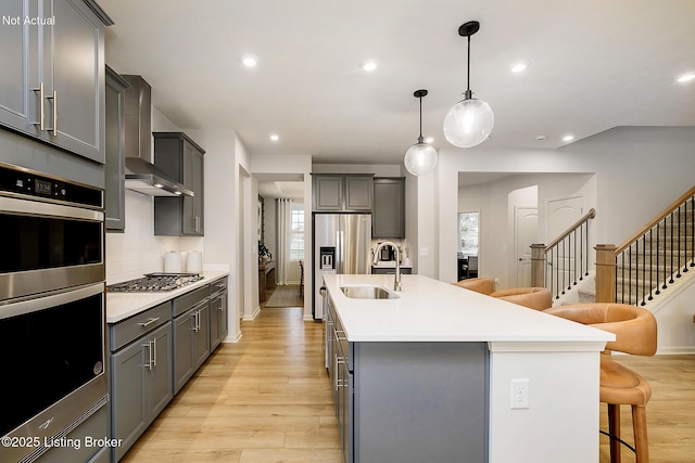 kitchen featuring a breakfast bar area, stainless steel appliances, gray cabinetry, wall chimney range hood, and a sink