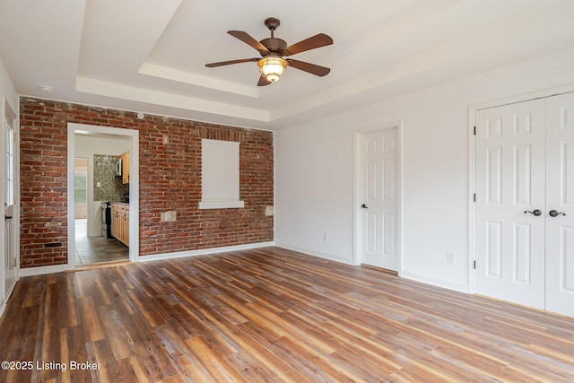 unfurnished bedroom with a raised ceiling, ensuite bathroom, brick wall, wood finished floors, and baseboards