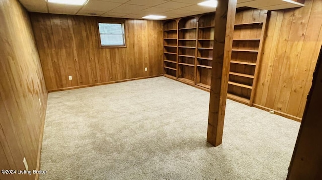 basement featuring a paneled ceiling, wooden walls, baseboards, and carpet flooring