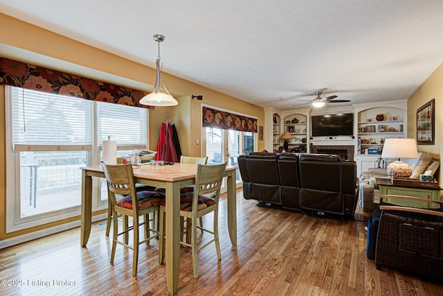 dining space with built in shelves, a fireplace, wood finished floors, and a ceiling fan