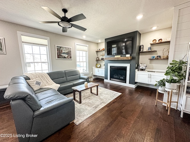 living area featuring a textured ceiling, ceiling fan, dark wood-style flooring, and a glass covered fireplace