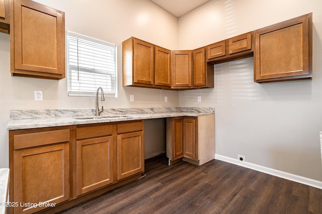 kitchen featuring light stone counters, dark wood-style floors, brown cabinetry, baseboards, and a sink