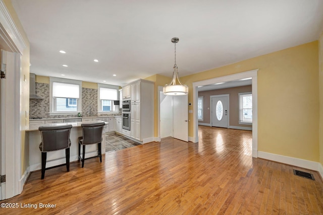 kitchen with tasteful backsplash, visible vents, wall chimney range hood, light wood-style flooring, and a peninsula