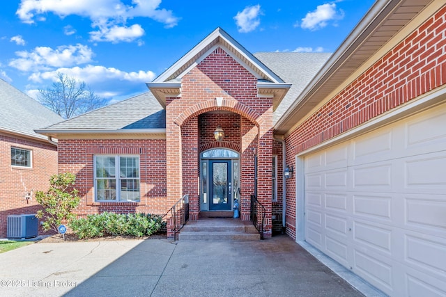 entrance to property featuring central air condition unit, driveway, an attached garage, a shingled roof, and brick siding