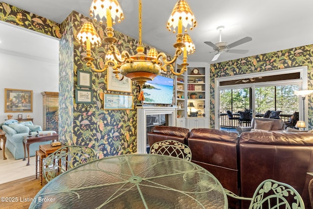 dining room with ceiling fan with notable chandelier, built in features, wood finished floors, a fireplace, and wallpapered walls