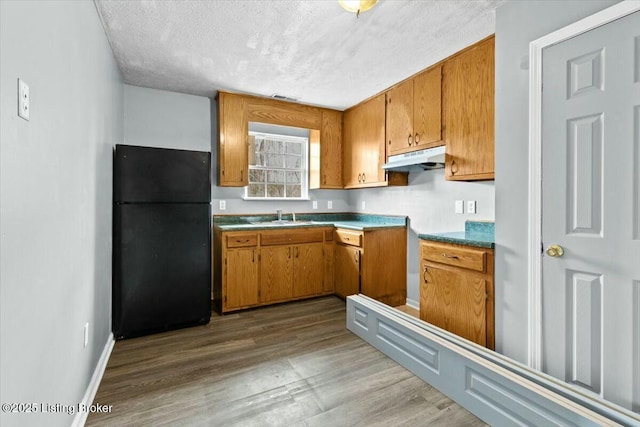 kitchen featuring brown cabinets, wood finished floors, freestanding refrigerator, under cabinet range hood, and a sink