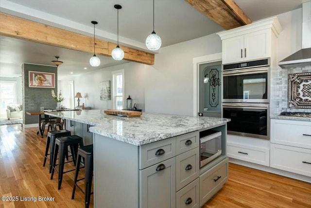 kitchen with light wood-type flooring, beam ceiling, backsplash, stainless steel appliances, and wall chimney range hood