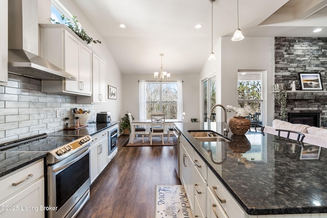 kitchen featuring dark wood finished floors, a sink, a stone fireplace, appliances with stainless steel finishes, and wall chimney exhaust hood