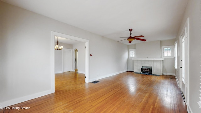 unfurnished living room with baseboards, ceiling fan with notable chandelier, a brick fireplace, and light wood finished floors