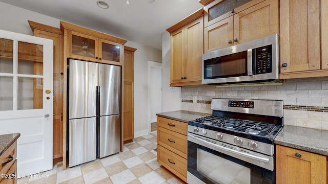 kitchen featuring tasteful backsplash, stainless steel appliances, dark stone counters, glass insert cabinets, and light floors