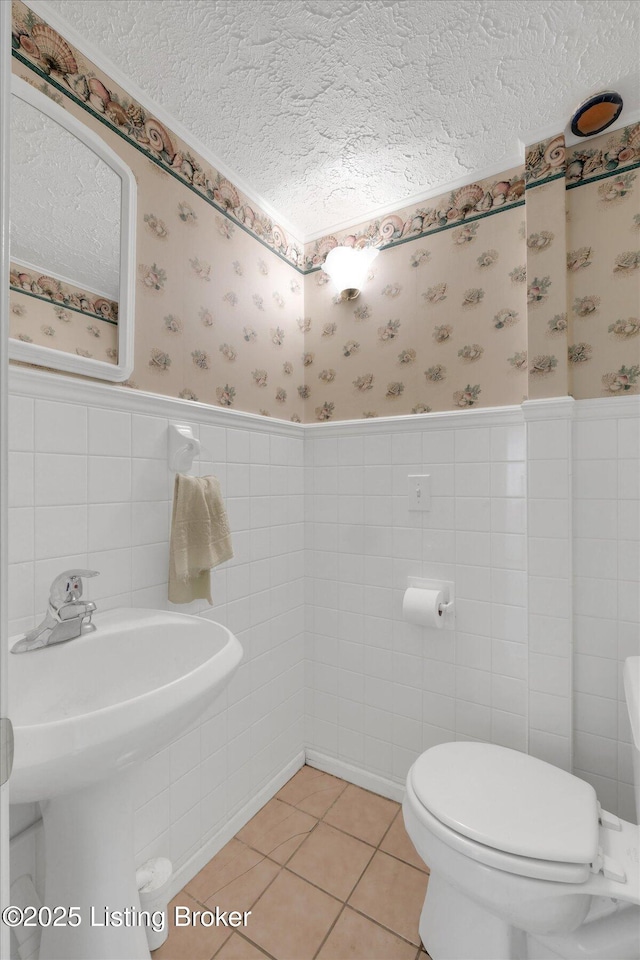 bathroom with a textured ceiling, wallpapered walls, wainscoting, tile patterned floors, and toilet