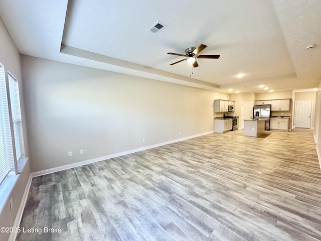 unfurnished living room with light wood finished floors, visible vents, baseboards, a tray ceiling, and a ceiling fan