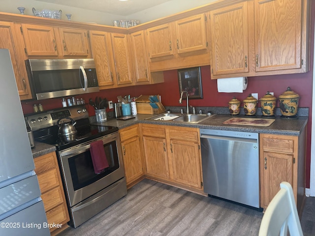 kitchen with brown cabinetry, light wood finished floors, a sink, stainless steel appliances, and dark countertops