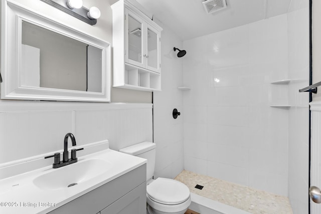 full bath with visible vents, a wainscoted wall, toilet, tiled shower, and vanity