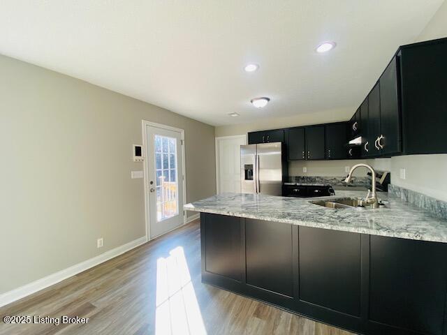 kitchen with dark cabinetry, light stone counters, a peninsula, a sink, and stainless steel refrigerator with ice dispenser