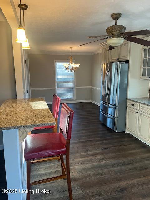 kitchen with light stone counters, ornamental molding, dark wood-type flooring, stainless steel refrigerator with ice dispenser, and ceiling fan with notable chandelier