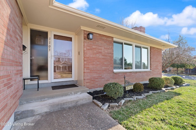 entrance to property with visible vents, a lawn, brick siding, and a chimney