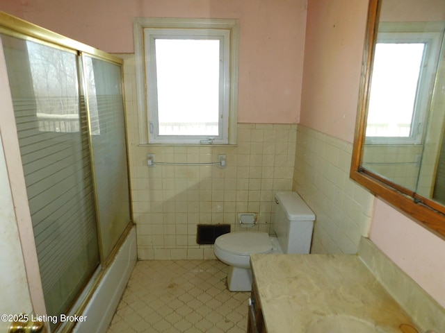 bathroom featuring a wainscoted wall, toilet, vanity, combined bath / shower with glass door, and tile walls