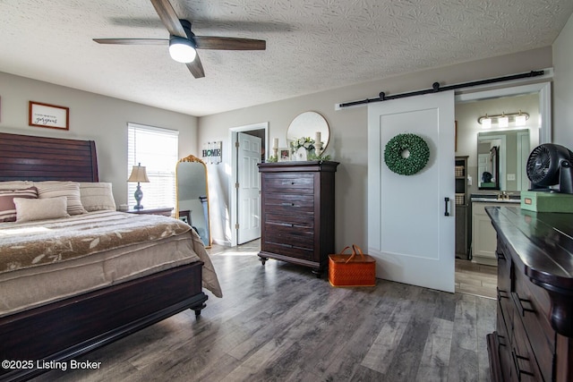 bedroom featuring a textured ceiling, ensuite bath, wood finished floors, a barn door, and ceiling fan