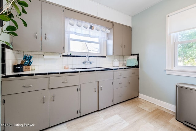 kitchen featuring tile countertops, baseboards, gray cabinetry, a sink, and backsplash