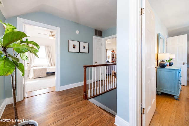 hallway with light wood-type flooring, visible vents, an upstairs landing, baseboards, and vaulted ceiling