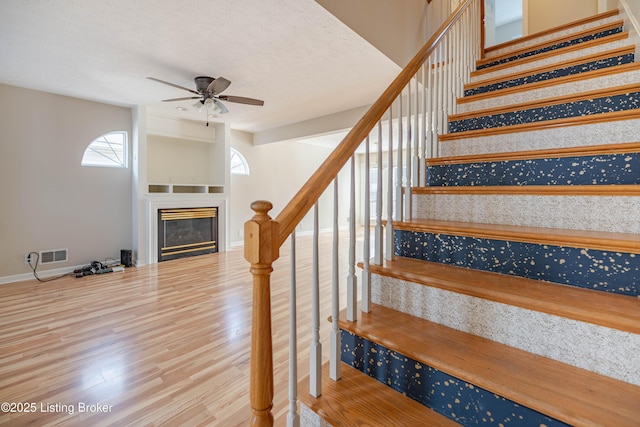 stairway with visible vents, baseboards, wood finished floors, a glass covered fireplace, and a ceiling fan