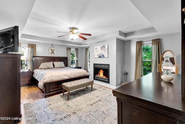 bedroom featuring multiple windows, a raised ceiling, a warm lit fireplace, and light wood finished floors