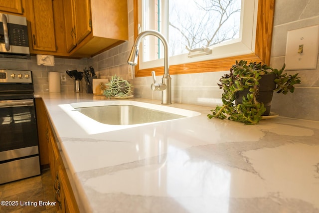 interior details with a sink, light stone counters, tasteful backsplash, appliances with stainless steel finishes, and brown cabinetry