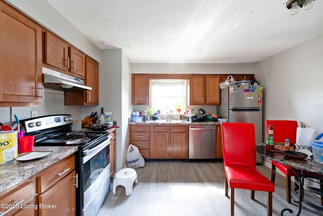 kitchen featuring light wood finished floors, under cabinet range hood, light stone counters, appliances with stainless steel finishes, and brown cabinetry