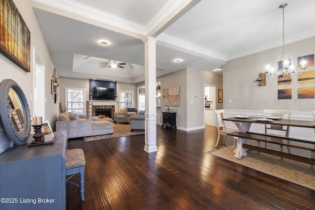 living room featuring a tiled fireplace, decorative columns, ceiling fan with notable chandelier, dark wood-style floors, and a raised ceiling