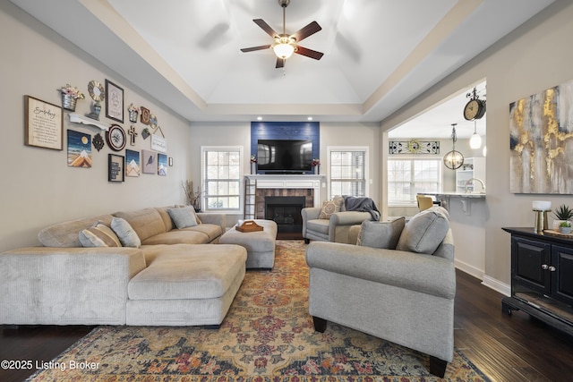 living room with baseboards, a fireplace, dark wood-style floors, a raised ceiling, and a ceiling fan