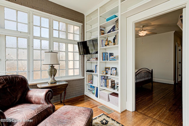 living area featuring a ceiling fan, brick wall, wood finished floors, and crown molding