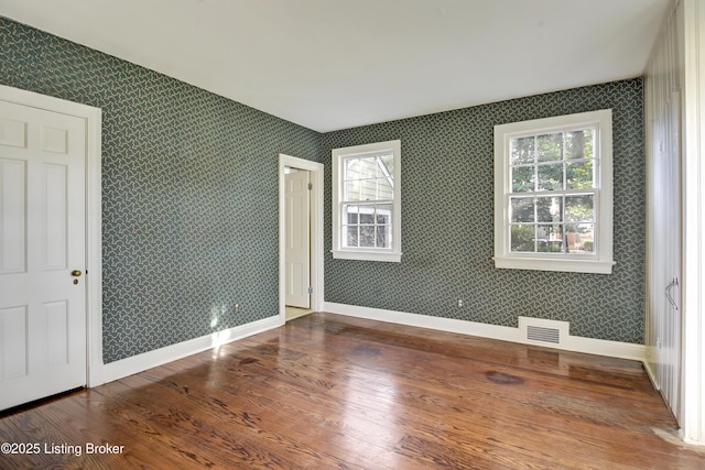 empty room featuring visible vents, wallpapered walls, baseboards, a wealth of natural light, and wood finished floors