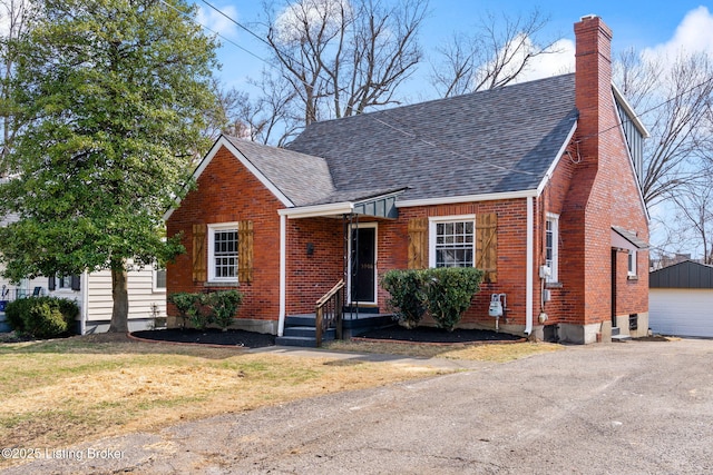 view of front facade featuring a front lawn, roof with shingles, an outdoor structure, brick siding, and a chimney