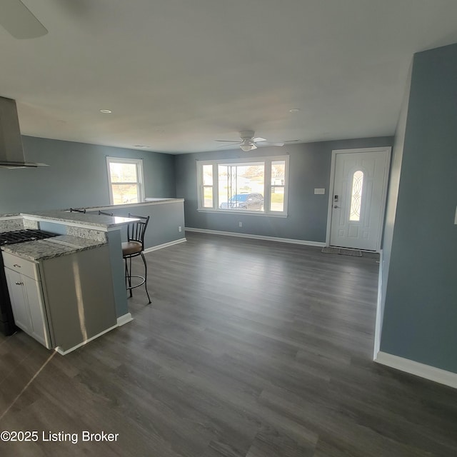 interior space featuring a ceiling fan, dark wood-style floors, a breakfast bar area, wall chimney exhaust hood, and baseboards