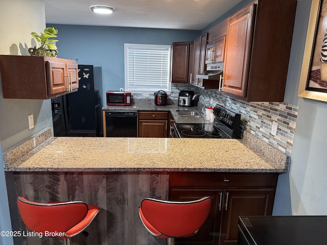 kitchen with black appliances, under cabinet range hood, a sink, backsplash, and light stone countertops