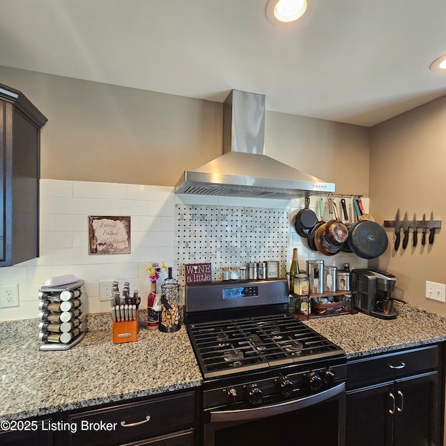 kitchen featuring backsplash, light stone countertops, stainless steel gas stove, wall chimney exhaust hood, and dark cabinets