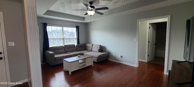 living area with ceiling fan, baseboards, a raised ceiling, and dark wood-style floors