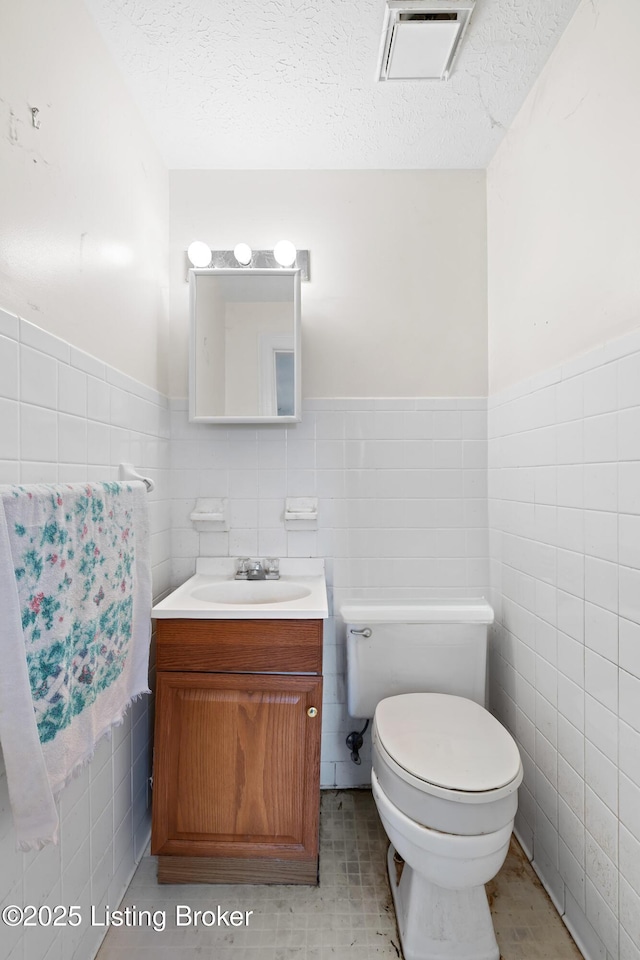 bathroom with visible vents, toilet, a textured ceiling, tile walls, and vanity