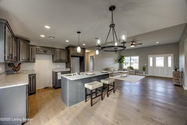 kitchen featuring light stone countertops, open floor plan, an island with sink, light wood-style flooring, and ceiling fan with notable chandelier