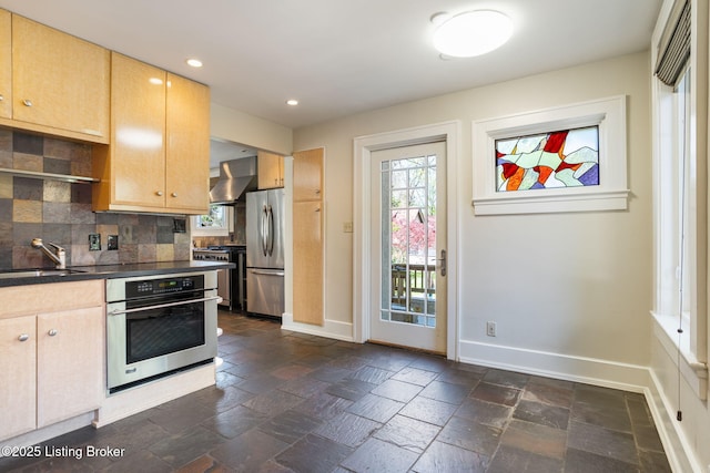 kitchen with baseboards, wall chimney exhaust hood, a sink, stone tile flooring, and stainless steel appliances